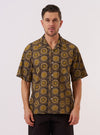A model wears the Olive Cape Block Print Camp Shirt against a plain white background. The shirt features an olive green block print pattern throughout, a classic camp collar, and a buttoned front.
