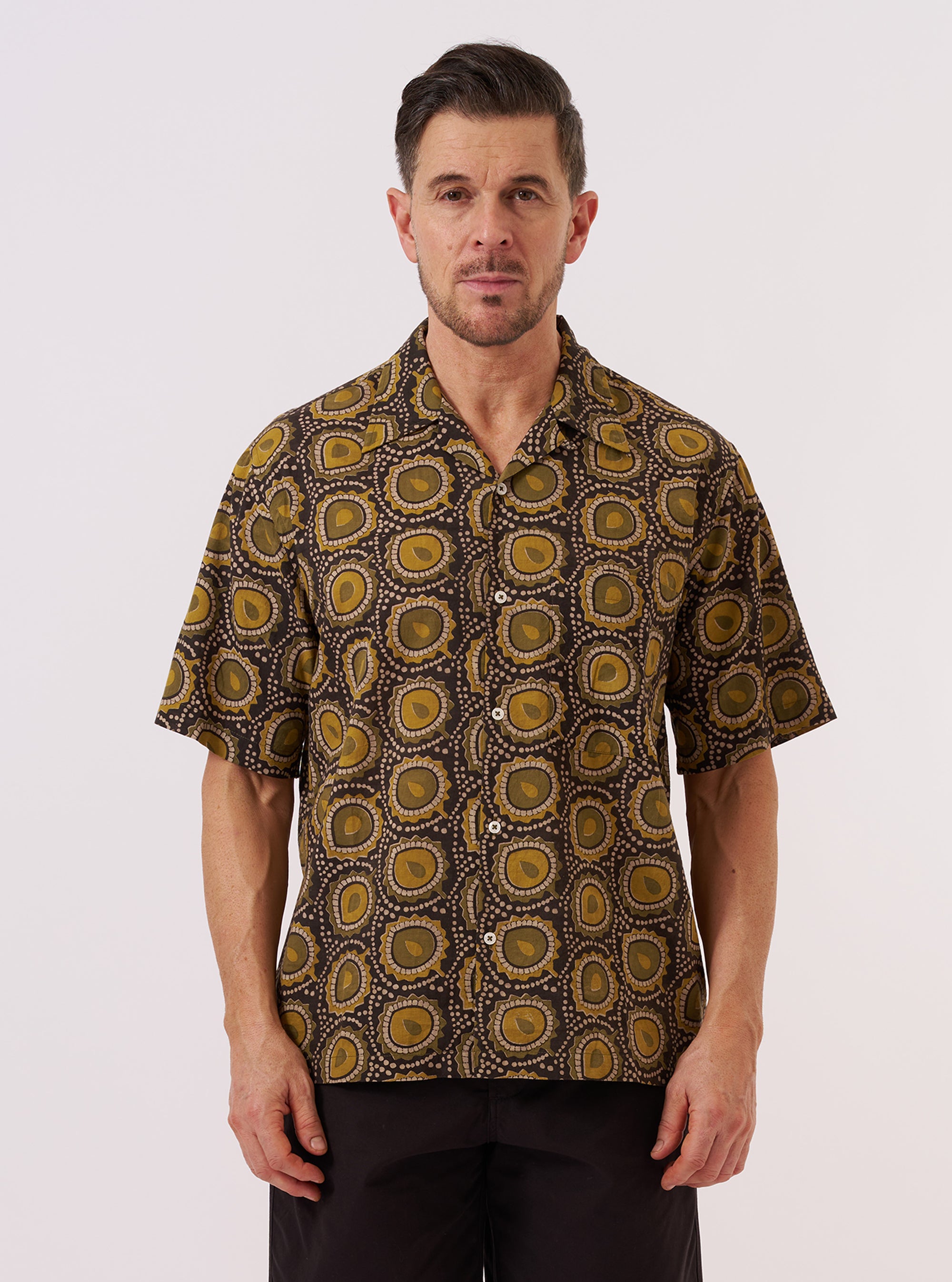 A model wears the Olive Cape Block Print Camp Shirt against a plain white background. The shirt features an olive green block print pattern throughout, a classic camp collar, and a buttoned front.