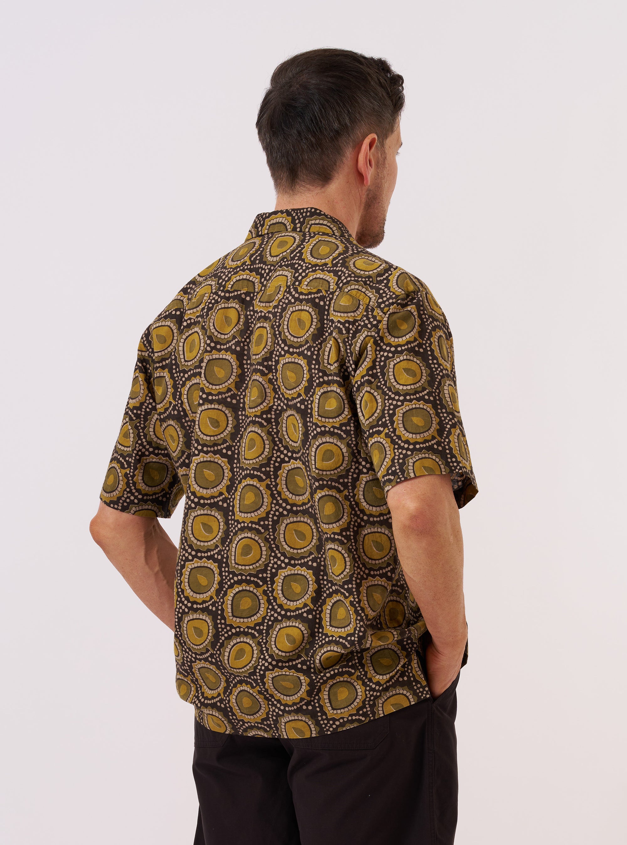 The Olive Cape Block Print Camp Shirt is worn by a model against a plain white background. The shirt is shown from the back, highlighting the back yoke with small pleats and the straight, loose fit of the short sleeves. The model wears the shirt untucked over dark trousers. No mannequin is present in the image.