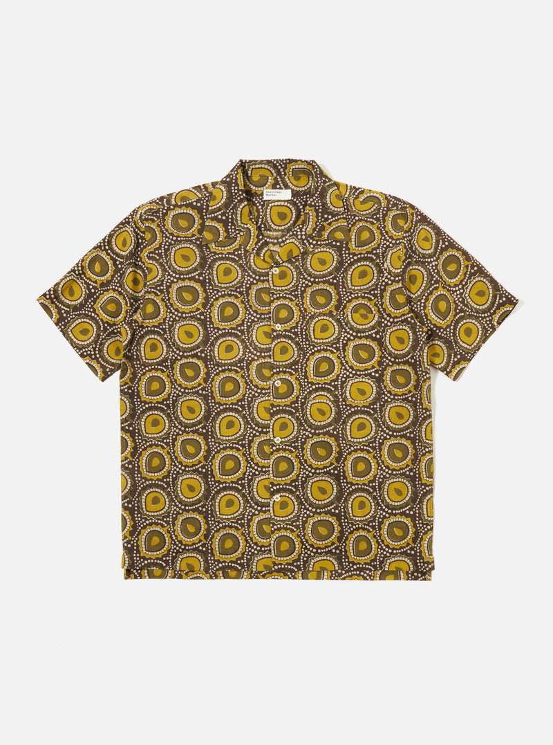 The Olive Cape Block Print Camp Shirt is shown as a flat lay against a white background. The shirt features a short-sleeved design with a camp collar, a single chest pocket, and a front button closure. The fabric is decorated with a block-printed pattern of white motifs on an olive green base.