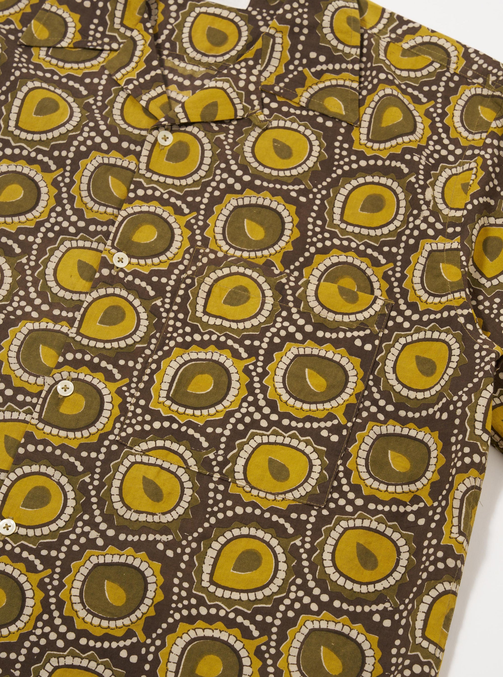 The Olive Cape Block Print Camp Shirt is shown from the back as a flat lay against a plain white background. The image highlights the straight hem, the back of the camp collar, and the intricate, repeating block print pattern featuring botanical motifs in shades of green and brown on an olive base.