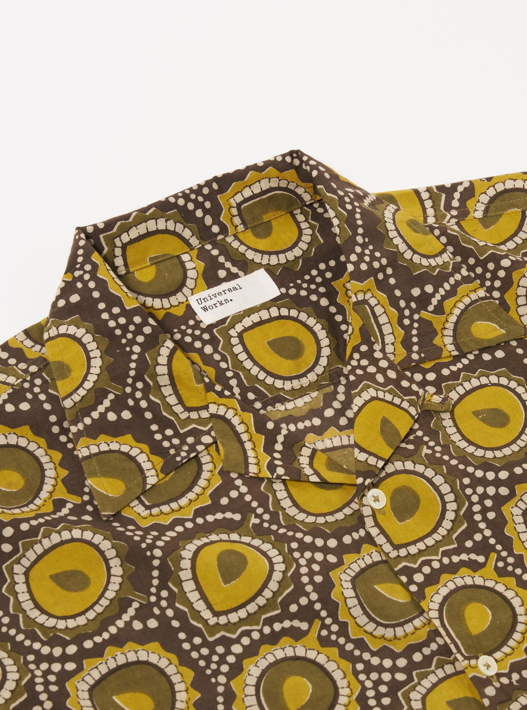 The Olive Cape Block Print Camp Shirt is shown in a detail view as a flat lay against a plain white background. The image highlights the side split hem, which is slightly longer at the back, and the loose fit of the short sleeve. The hand-printed olive pattern is clearly visible on the cotton fabric.