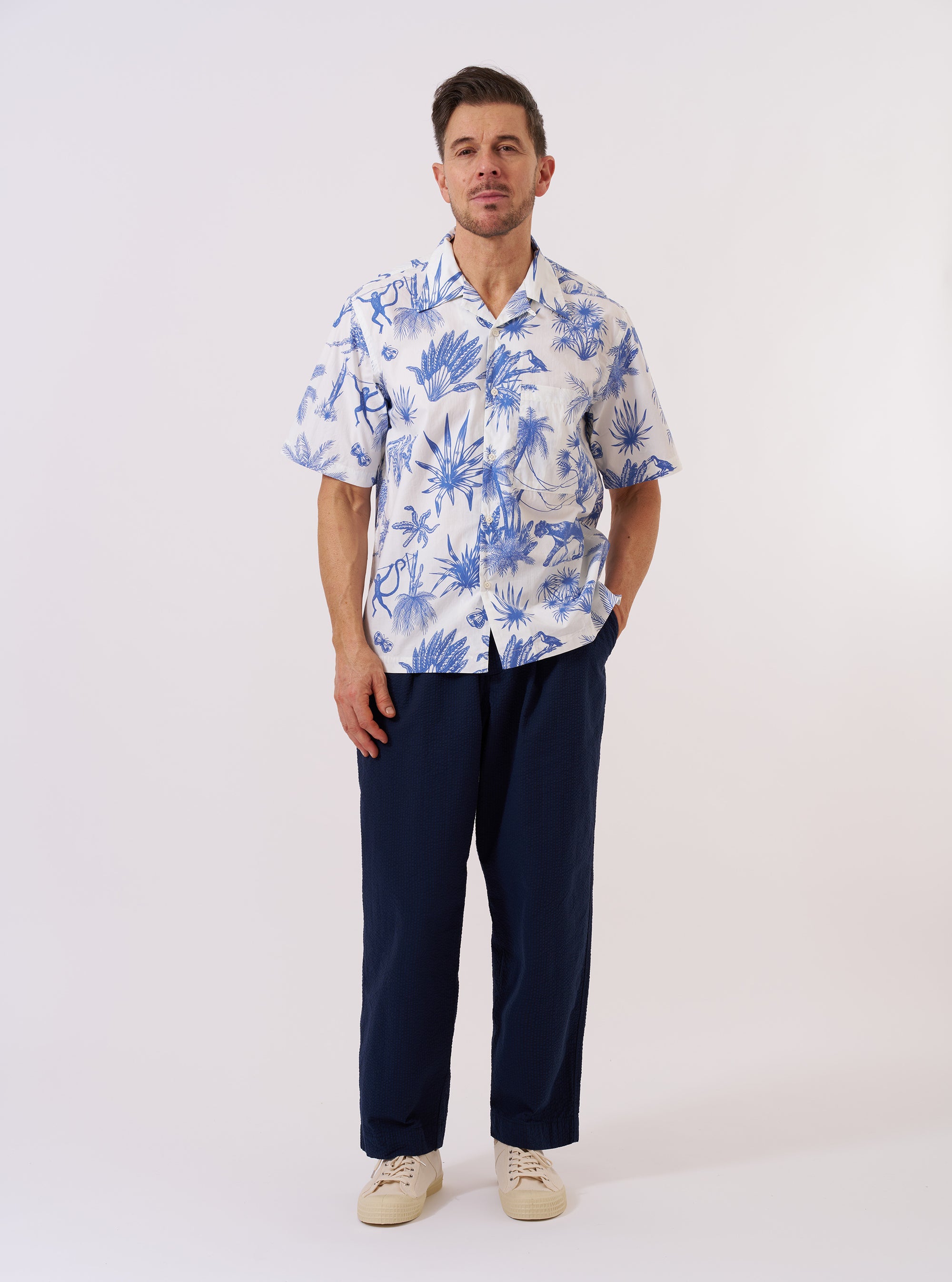 The White Jungle Toile Poplin Camp Shirt is shown as a flat lay against a white background. The shirt features a camp collar, a single chest patch pocket, and a central button-down front. The fabric is a white cotton poplin decorated with a detailed jungle toile print featuring animals and tropical foliage in a dark ink.