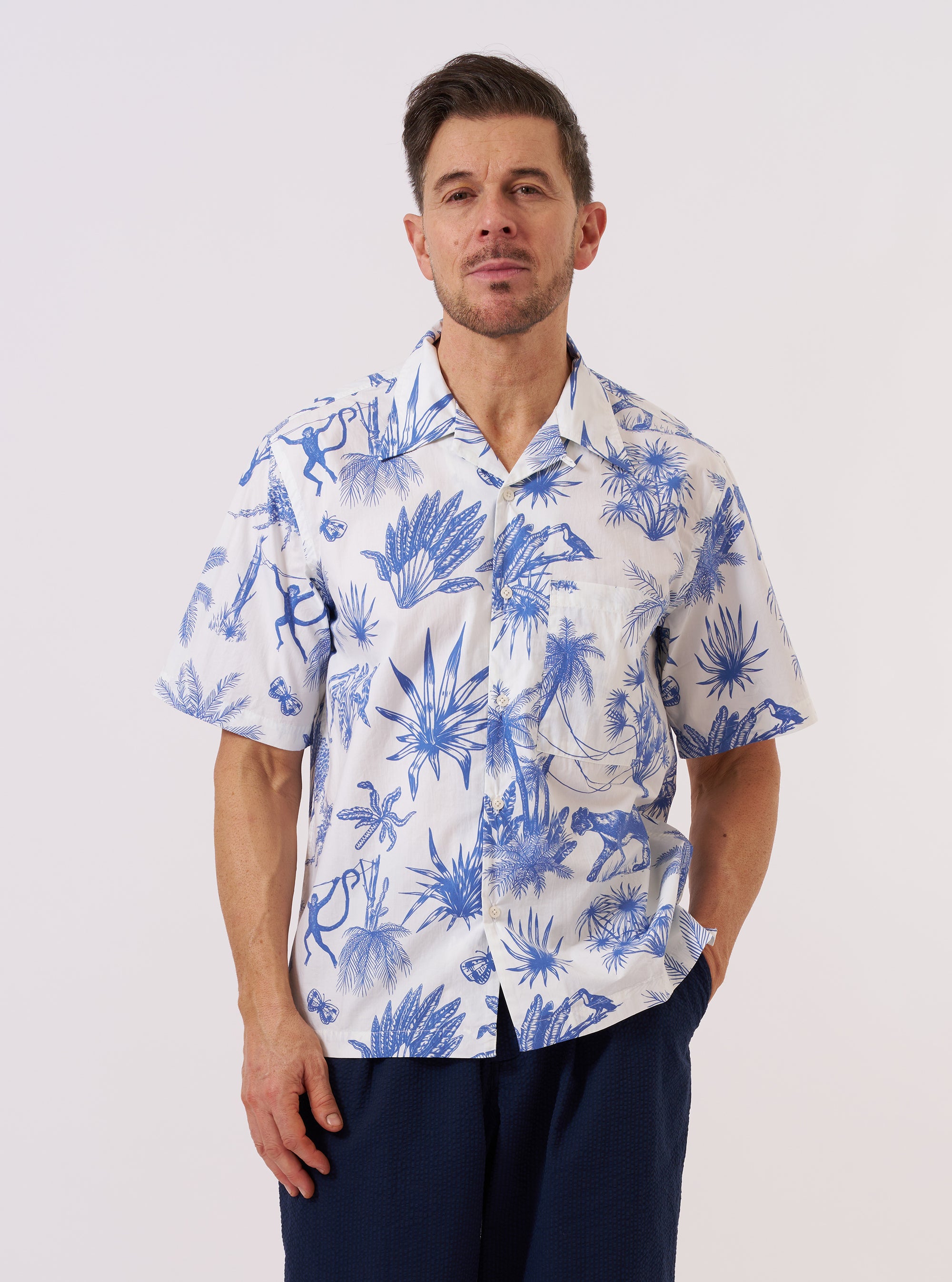 A model wears the White Jungle Toile Poplin Camp Shirt against a plain white background. The shirt features an all-over dark jungle toile print on a white base, a relaxed camp collar, and a central button-down front. The model is shown from the chest up with the shirt worn open over a white T-shirt.