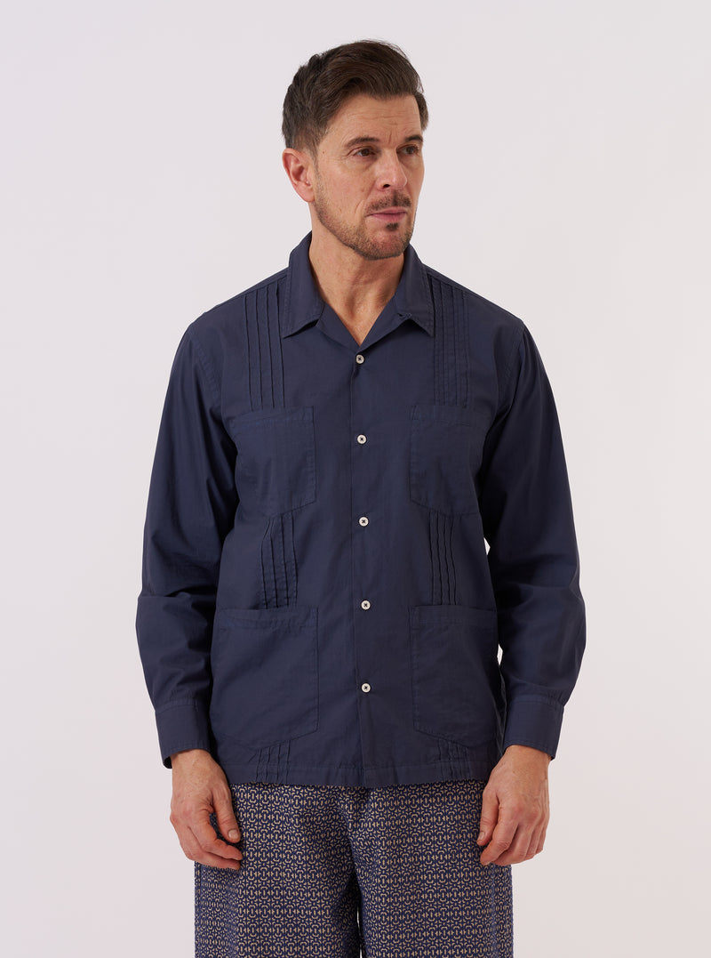 A model wears the Navy Poplin Dwight Overshirt over a light-colored top against a plain, neutral background. The image highlights the garment's distinctive vertical pleats running down the front, four large patch pockets, and a classic Cuban collar. The shirt is shown in a navy blue poplin fabric with a straight hem.