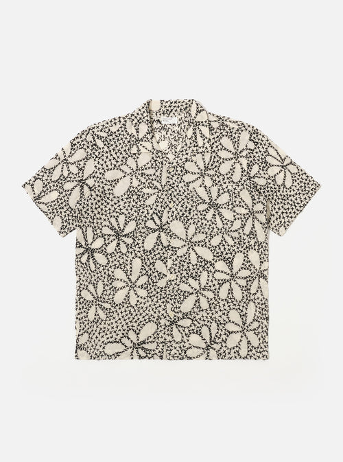 The Ecru/Black Kantha Flower Road Shirt is shown as a flat lay against a white background. The short-sleeve shirt features a camp collar, a central button-down front, and a single patch pocket on the left chest. The ecru fabric is detailed with an all-over black floral embroidery pattern.