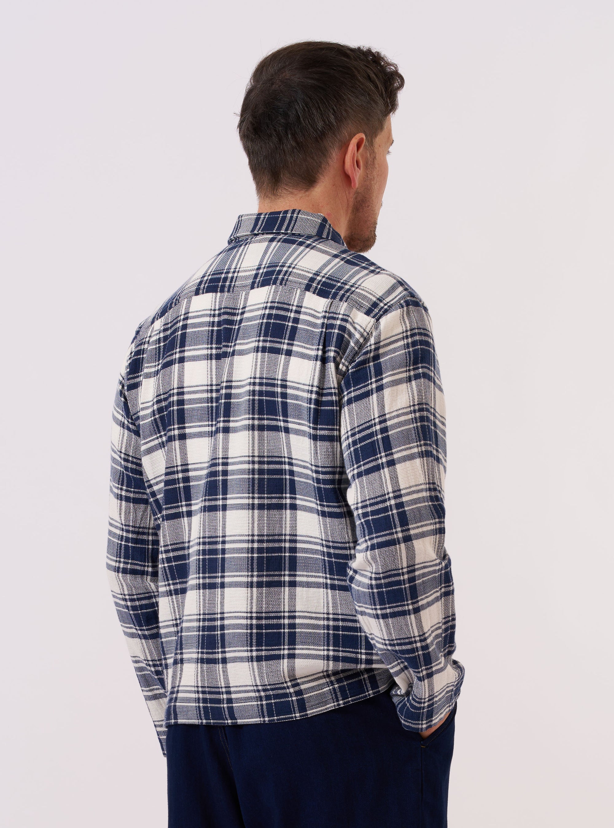 A model wears the Navy Texture Plaid Zip Shirt over a light-colored top, shown from a rear three-quarter angle against a plain white background. The shirt features a relaxed fit, a classic pointed collar, and a distinct textured navy blue plaid pattern throughout. The image highlights the fabric's drape and the garment's straight hem.