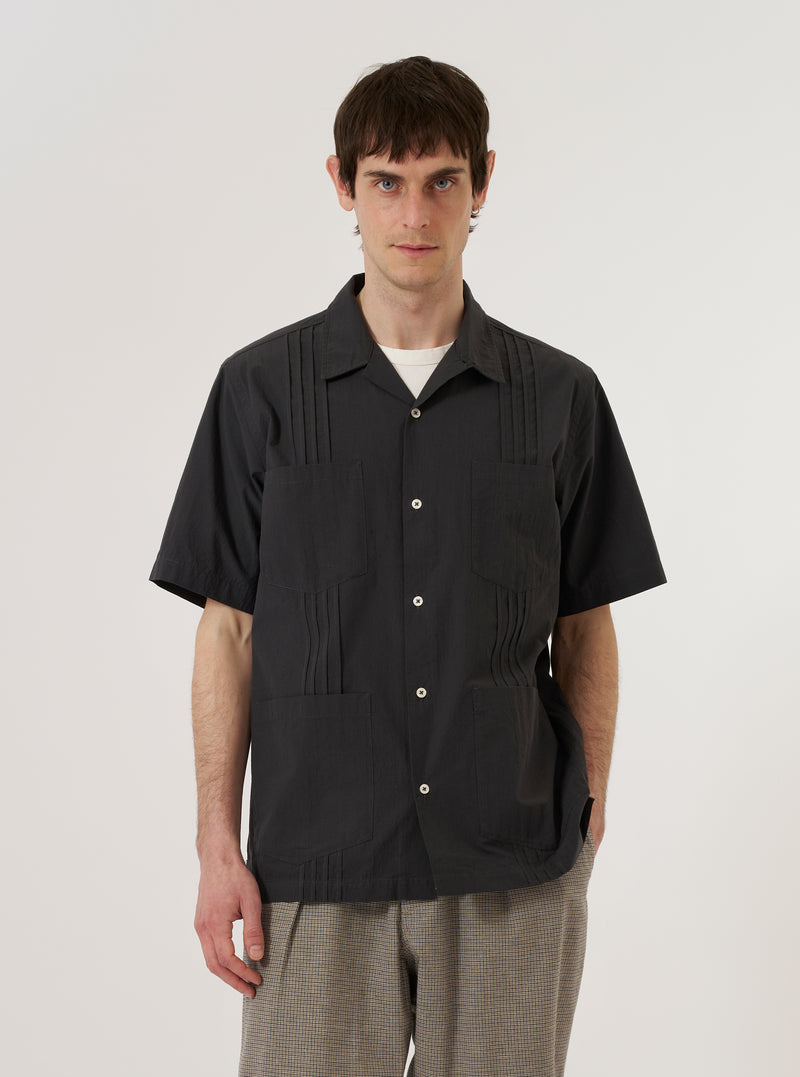 A model wears the Black Tech Cotton S/S Dwight Overshirt against a plain white background. The short-sleeved overshirt features a camp collar, a button-up front, and two large utility-style patch pockets on the chest. The model wears the garment unbuttoned over a white t-shirt, showcasing its relaxed fit and the smooth, technical texture of the black cotton fabric.
