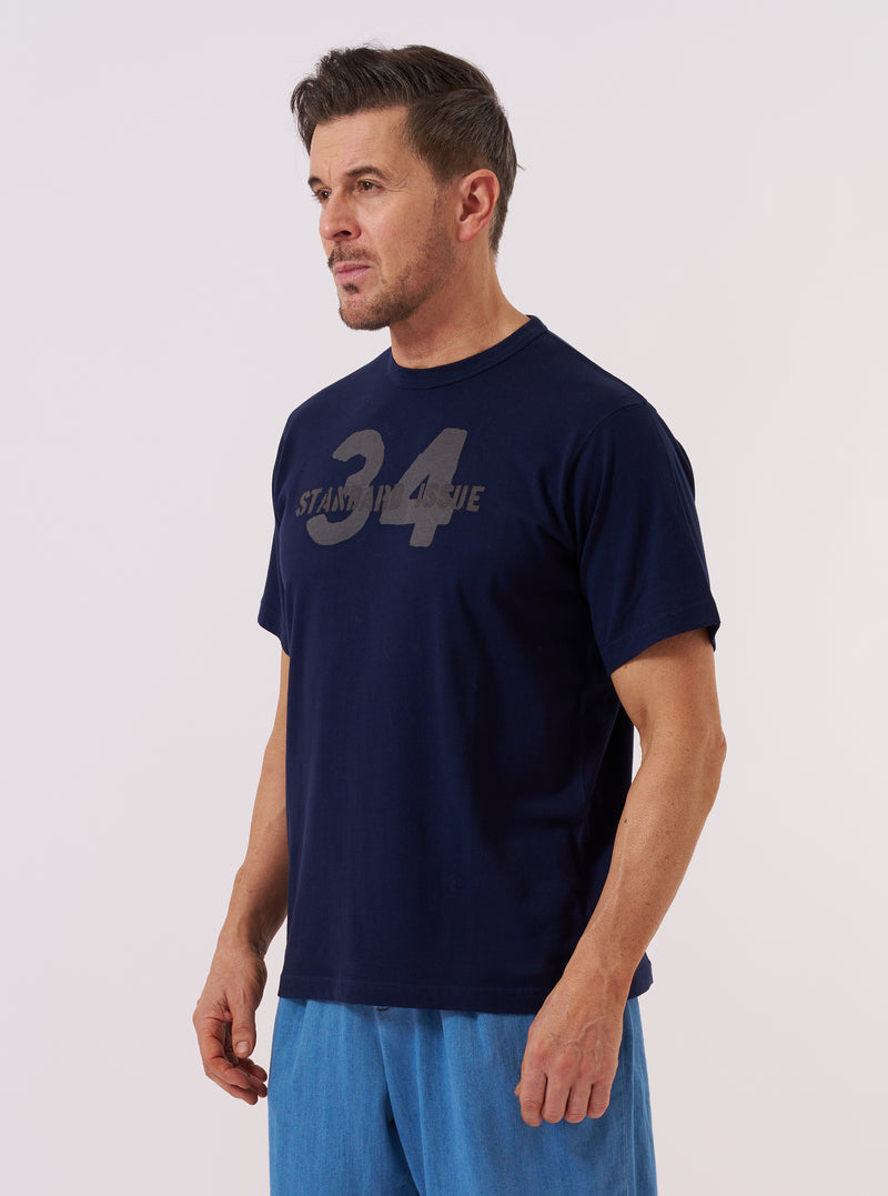 A model wears the Navy Single Jersey 34 Printed Core Tee against a plain white background. The short-sleeve t-shirt features a classic crew neckline and a prominent graphic print on the chest. The garment is constructed from a dark navy single jersey cotton fabric.