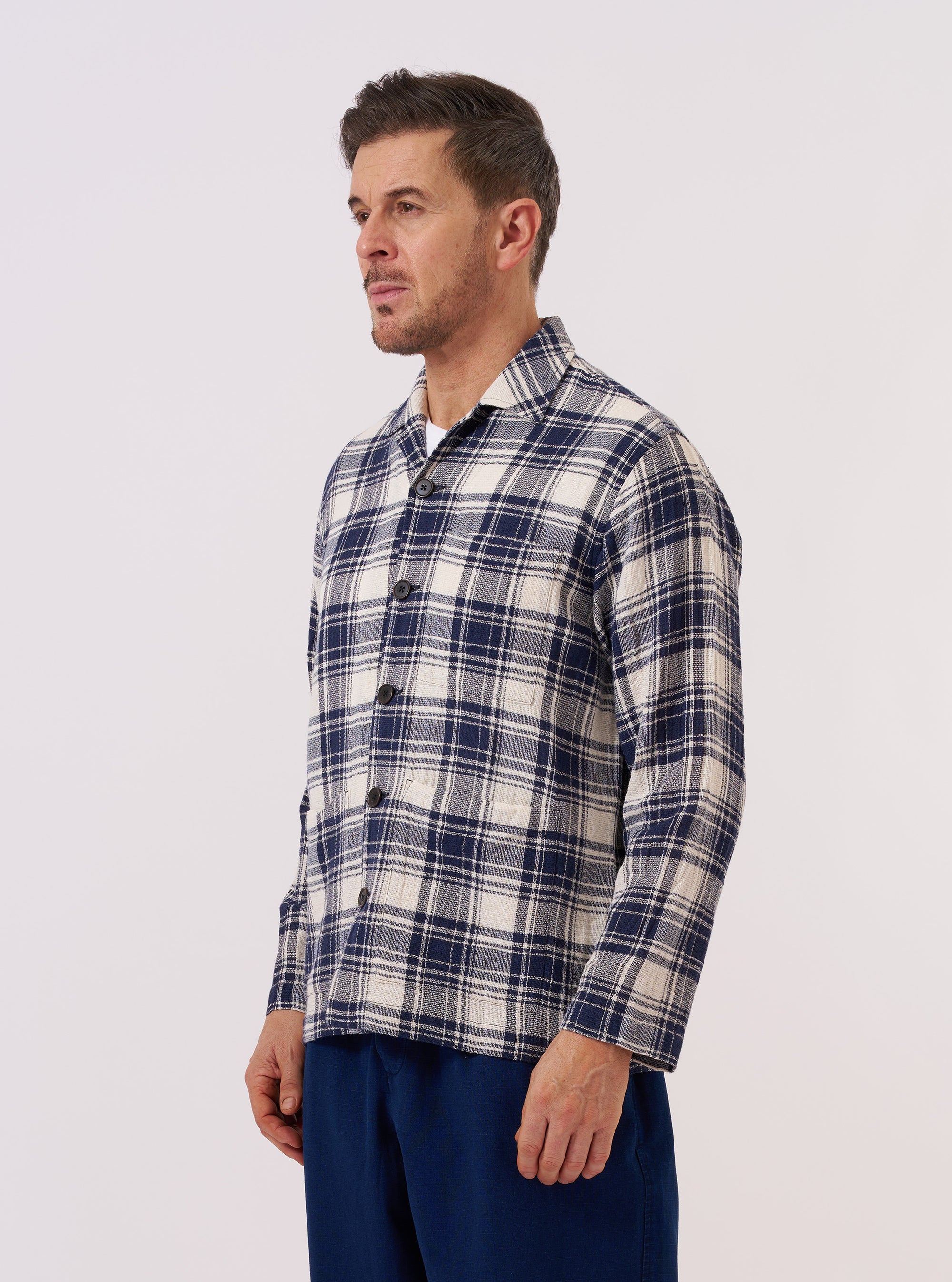 A model wears the Navy Texture Plaid Cancun Jacket over a white t-shirt and light-colored trousers against a plain, light grey background. The jacket is a boxy, overshirt-style garment featuring a notched collar, a three-button front closure, one chest pocket, and two large hip pockets. The navy blue cotton fabric has a distinct textured plaid pattern. The model is standing with one hand in their trouser pocket, showing the jacket's relaxed fit and low hip length.