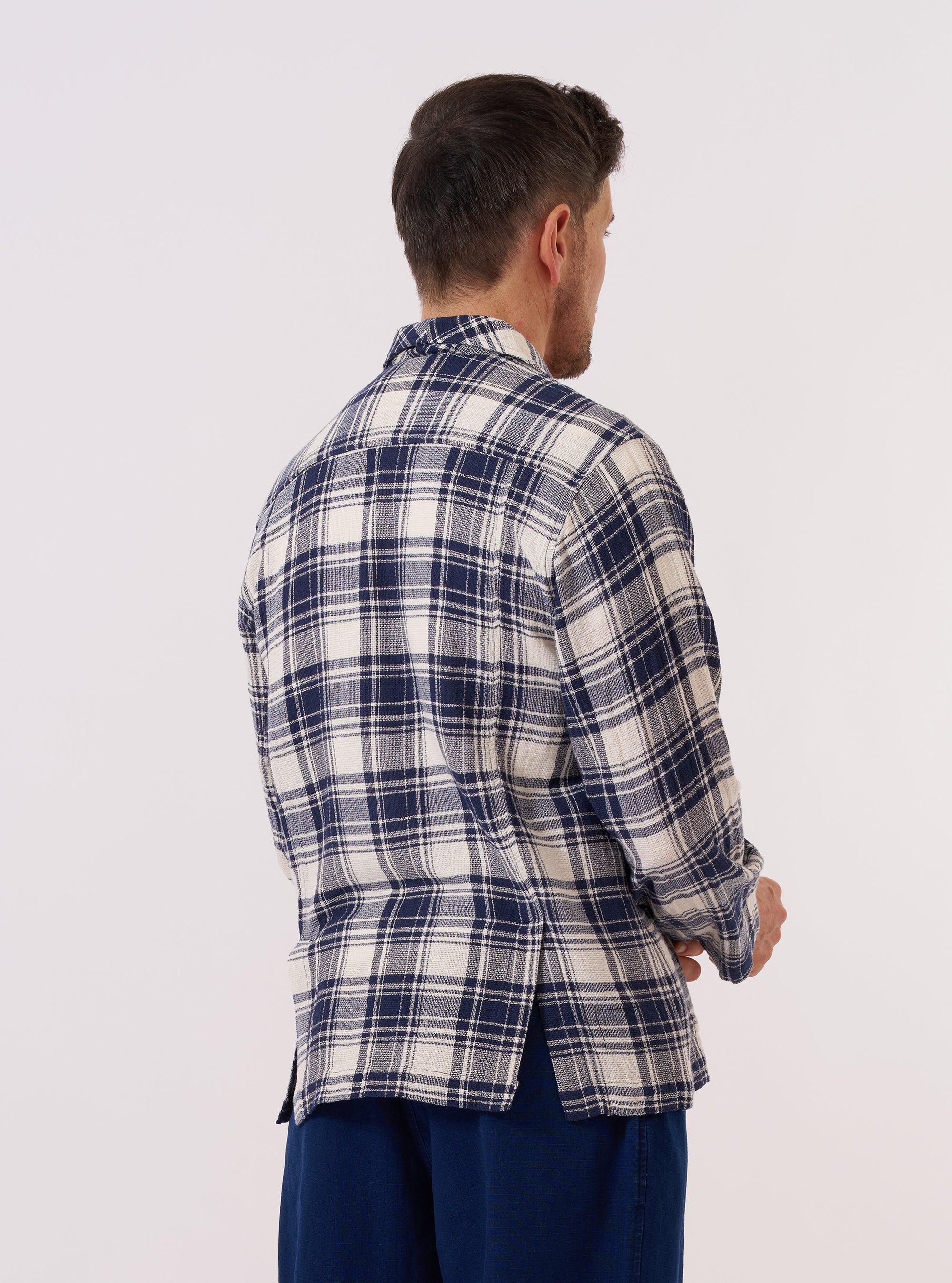 A model wears the Navy Texture Plaid Cancun Jacket against a plain, light-colored background. The jacket is styled as a smarter overshirt featuring a notched collar, a single chest pocket, and two lower hip pockets. The navy cotton fabric has a distinct textured plaid pattern. The model is shown from the front, highlighting the boxy fit and button-down construction of the garment.