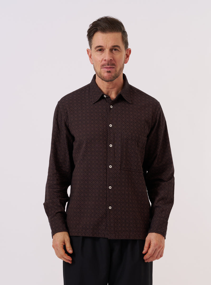 A model wears the Brown Diamond Dobby Lucky Shirt over a light-colored t-shirt, standing against a plain, neutral background. The shirt features a short-sleeve design, a classic collar, and a central button closure. The brown cotton fabric is characterized by a visible, textured diamond dobby geometric pattern.