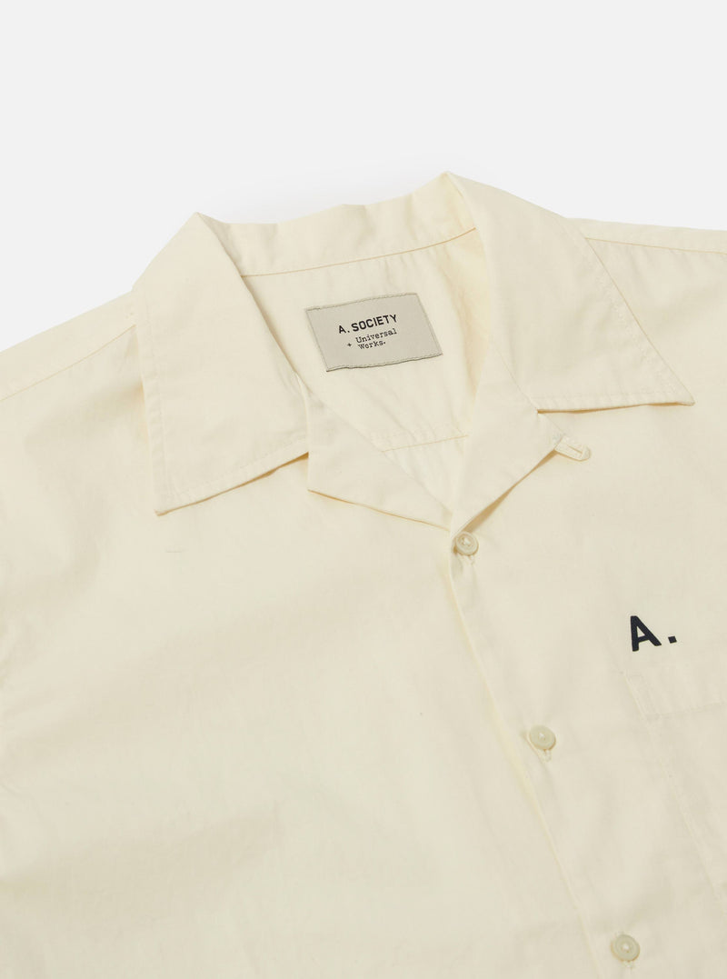 Close-up of the A. SOCIETY x Universal Works Ecru Broadcloth Camp Shirt on a white background. The shirt is ecru-colored with a camp collar and button-up front. 'A.' is embroidered on the pocket.