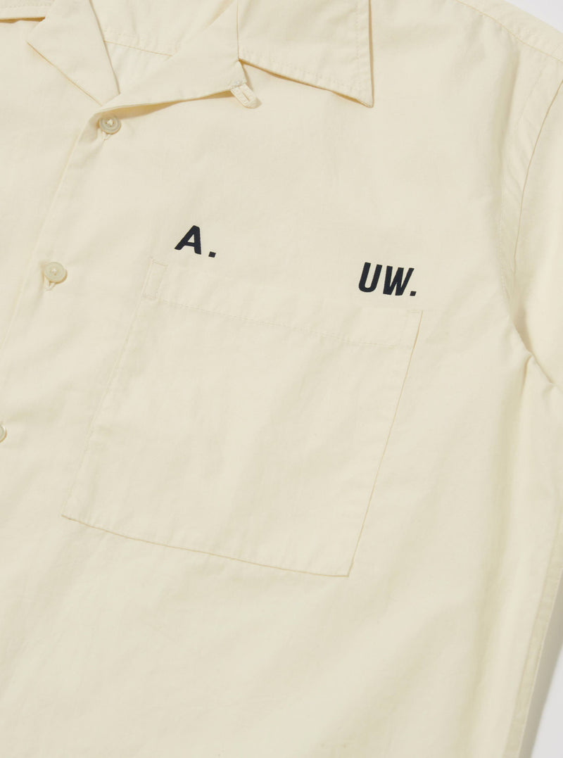 Close-up of the A. SOCIETY x Universal Works Ecru Broadcloth Camp Shirt. The ecru shirt features a chest pocket and black lettering. Shot on a plain white background.
