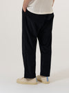 A model wears the Black Organic Fine Cord Oxford Pant, shown from the waist down against a plain, neutral background. The trousers feature a wide, loose leg with a single pleat front and are made from a black fine-wale corduroy fabric. The elasticated waistband with an external drawstring is clearly visible, and the model is standing with one hand in a front pocket, highlighting the garment's relaxed fit.