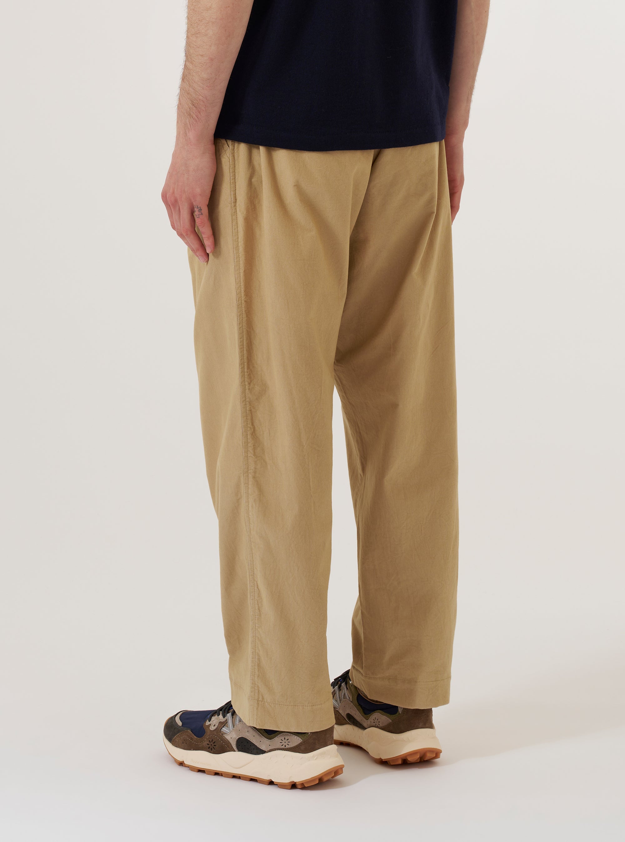 The Sand Organic Fine Cord Oxford Pant is worn by a model and shown from a rear three-quarter angle against a plain white background. The image highlights the relaxed, wide-leg fit of the trousers and the fine-ribbed texture of the sand-colored organic cotton corduroy. One rear pocket with a buttoned flap is visible. The model is wearing the pants with white socks and dark shoes.