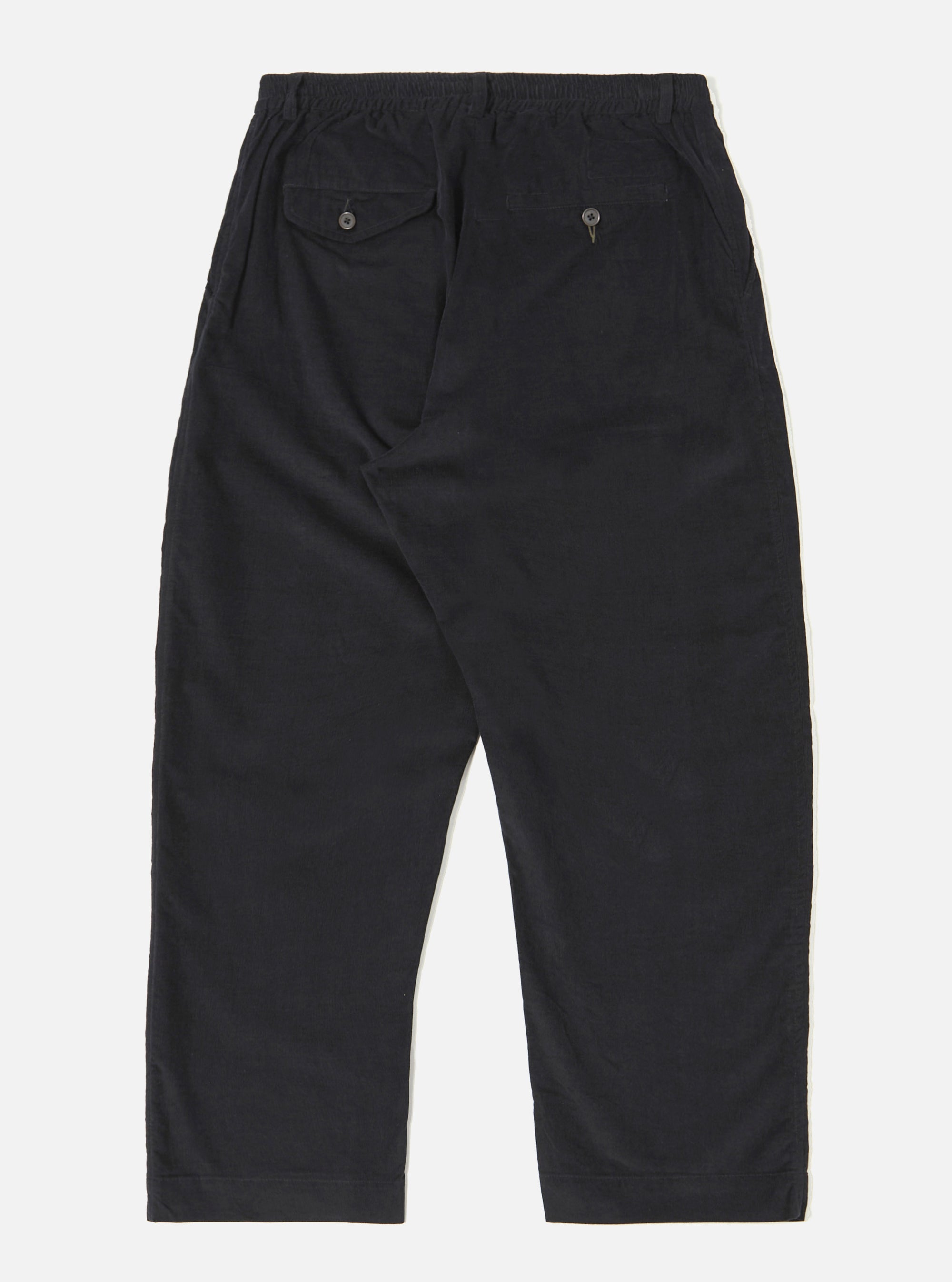 The Black Organic Fine Cord Oxford Pant is shown from the back as a flat lay against a plain white background. The image highlights the two rear pockets, one with a buttoned flap and one button jetted, as well as the elasticated waistband. The black organic cotton fabric has a visible fine corduroy texture.