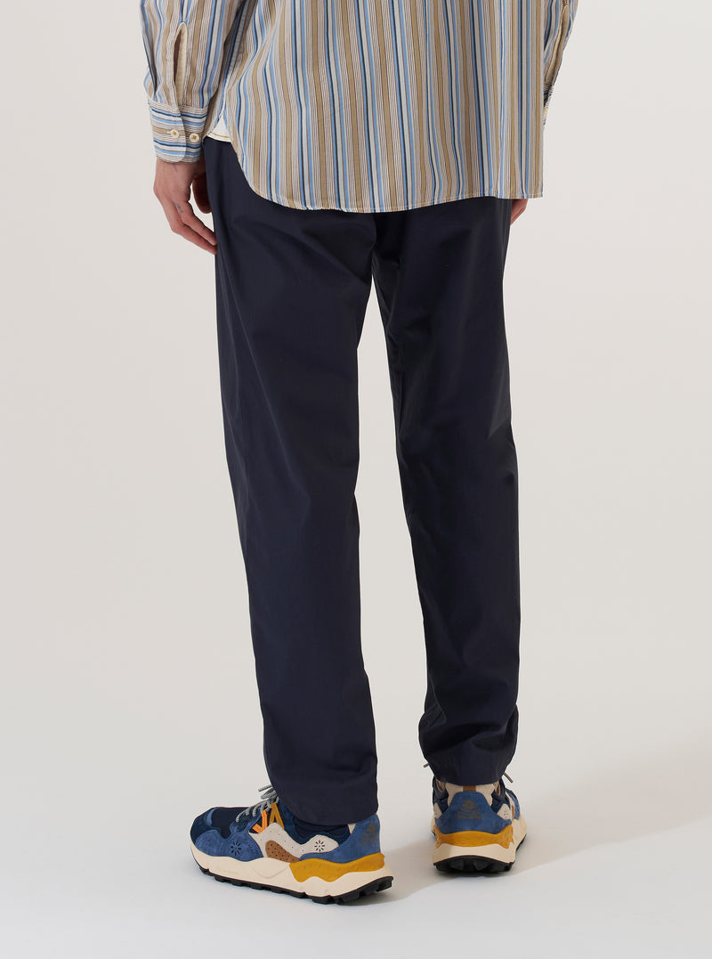 A model is wearing the Navy Peached Poplin Military Chino, shown from the waist down from a rear angle against a plain white background. The trousers feature a straight-leg silhouette and a rear flap pocket with a button closure. The navy blue fabric has a smooth, matte peached poplin finish.