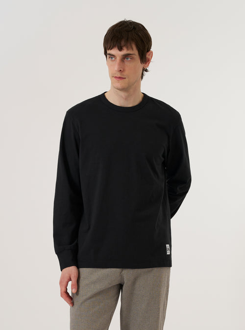 The Black Recycled/Bio Jersey Henry L/S Tee is worn by a model against a plain white background. The image, captured from the back at a slight angle, highlights the garment's relaxed fit, long sleeves, and clean neckline. The black jersey fabric has a smooth, matte finish. No mannequin is present in the image.