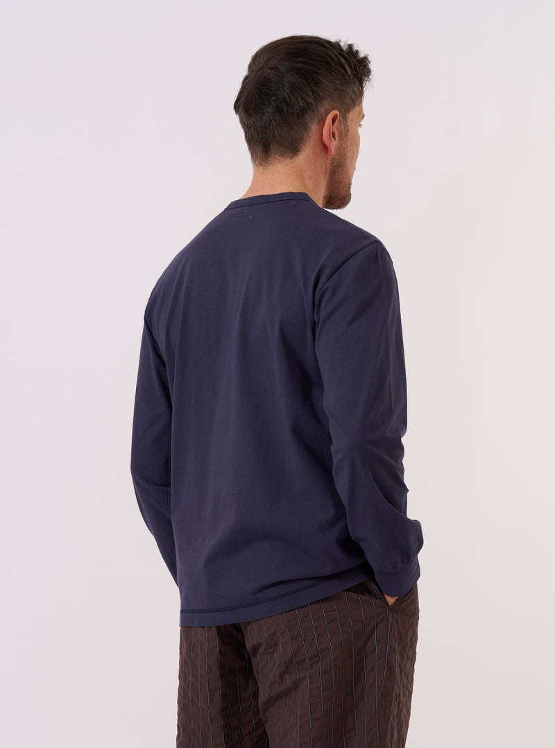 A model wears the Navy Recycled/Bio Jersey Henry L/S Tee, shown from the back against a plain white background. The long-sleeved garment features a relaxed fit and a straight hem, highlighting the soft texture and deep blue color of the recycled and organic cotton jersey fabric. No other accessories are prominent in this rear-view studio shot.