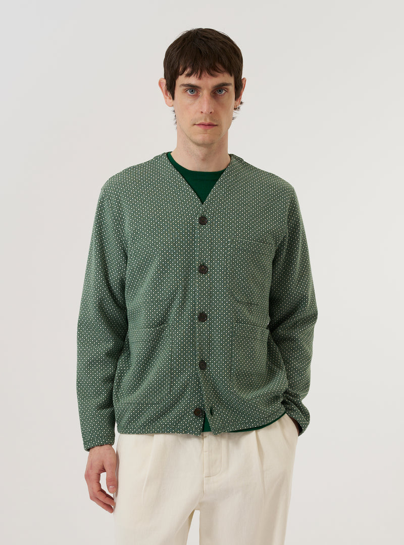 A model wears the Green Argyll Jersey Cardigan against a plain white background. The cardigan features an all-over tonal green Argyll diamond pattern, a five-button front closure, and ribbed detailing at the V-neckline, cuffs, and hem. The model is shown from the front, wearing the garment over a light-colored t-shirt.