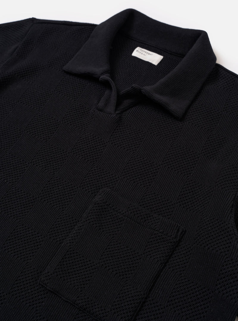 The Black Fantasy Checkerboard Vacation Polo is shown in a close-up detail shot as a flat lay against a plain white background. The image focuses on the upper portion of the garment, highlighting the open-neck design with a short placket, a single-piece collar, and a single patch chest pocket. The organic cotton fabric features a prominent black and grey fantasy checkerboard pattern.