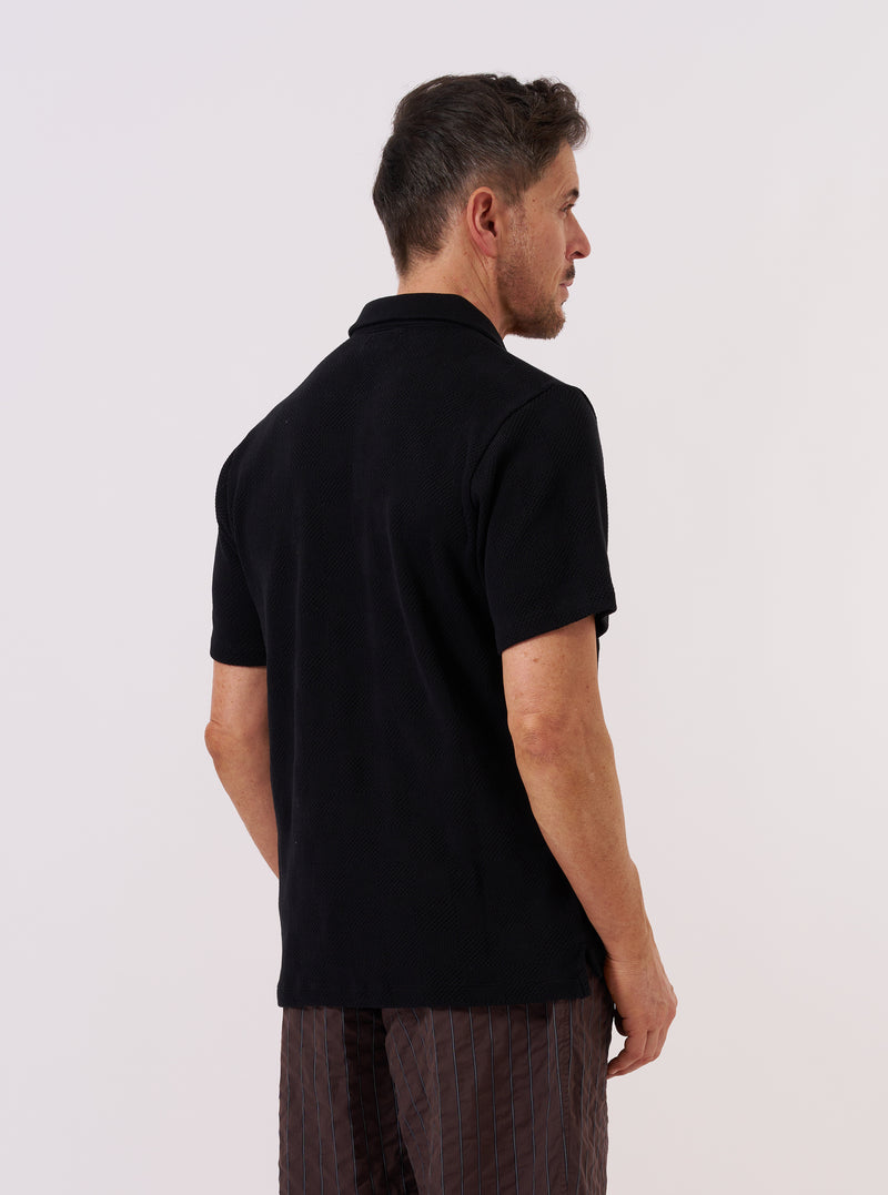 A model wears the Black Fantasy Checkerboard Vacation Polo, shown from a side angle against a plain, neutral studio background. The polo features an all-over black and grey checkerboard pattern, a relaxed fit, and a classic vacation collar. The textured organic cotton fabric and the short sleeves are prominent features in the image.