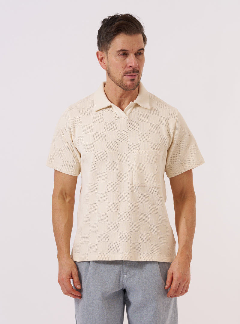 A model wears the Ecru Fantasy Checkerboard Vacation Polo against a plain white background. The polo features an all-over textured checkerboard pattern, a classic spread collar, and short sleeves. The garment is shown in a front view, highlighting the ecru organic cotton fabric and its distinct patterned weave.