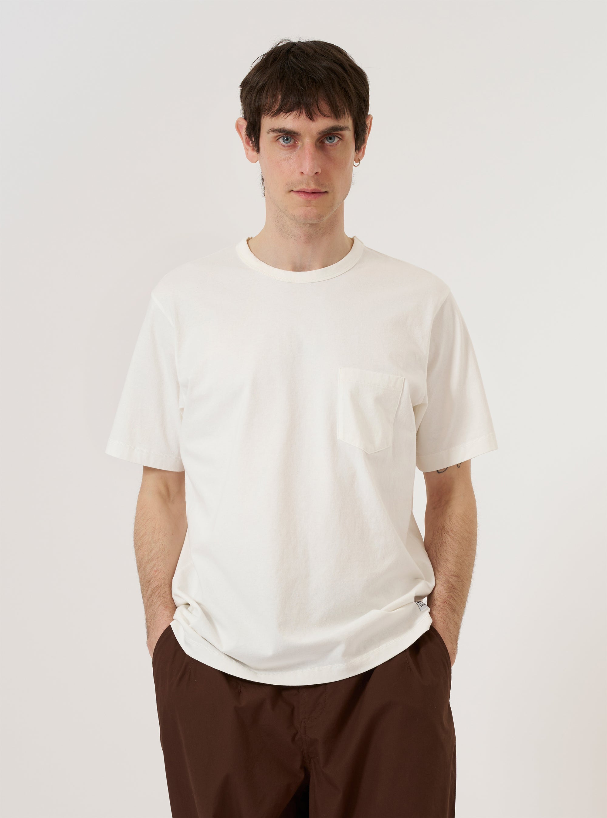 A model wears the Antique White Recycled/Bio Jersey Core Pocket Tee, standing against a plain white background. The T-shirt features a classic crew neck, short sleeves, and a single patch pocket on the left chest. The garment is made from a solid antique white jersey fabric with a smooth finish.