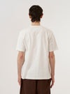 A model is wearing the Antique White Recycled/Bio Jersey Core Pocket Tee, shown from the chest down against a plain white background. The model is standing with one hand in their pocket, highlighting the relaxed fit of the t-shirt and the single patch pocket on the left chest. The fabric is a solid antique white jersey with a smooth finish.