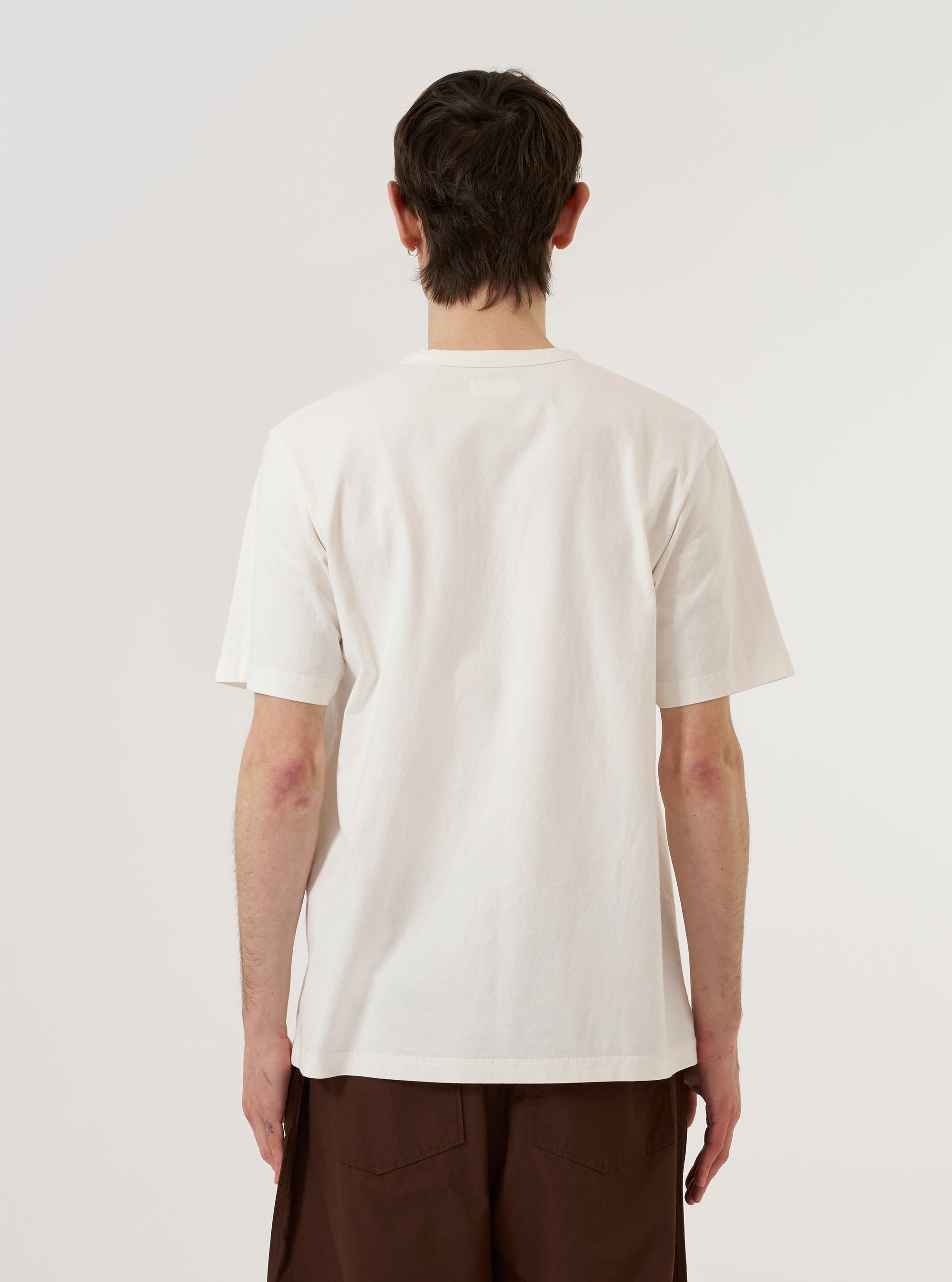 A model is wearing the Antique White Recycled/Bio Jersey Core Pocket Tee, shown from the chest down against a plain white background. The model is standing with one hand in their pocket, highlighting the relaxed fit of the t-shirt and the single patch pocket on the left chest. The fabric is a solid antique white jersey with a smooth finish.