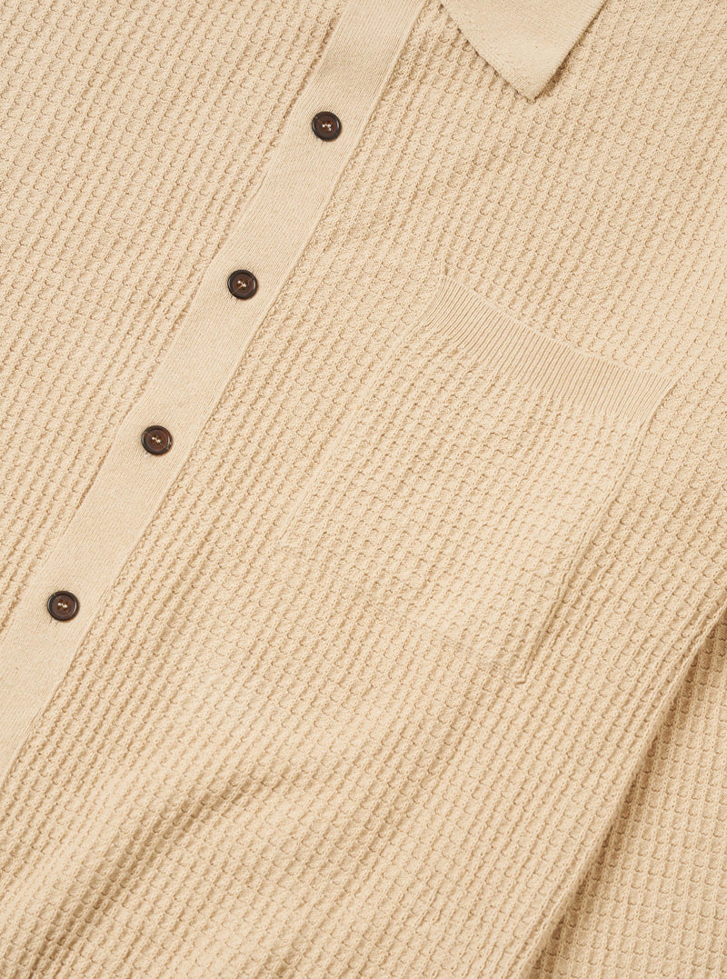 The Sand Eco Cotton Knit Jacket is shown from the back, worn by a model against a plain white background. The jacket features a clean, straight hem and a simple knit texture in a light sand color. The model is also wearing dark trousers, and the focus is on the fit and drape of the garment across the shoulders and back.