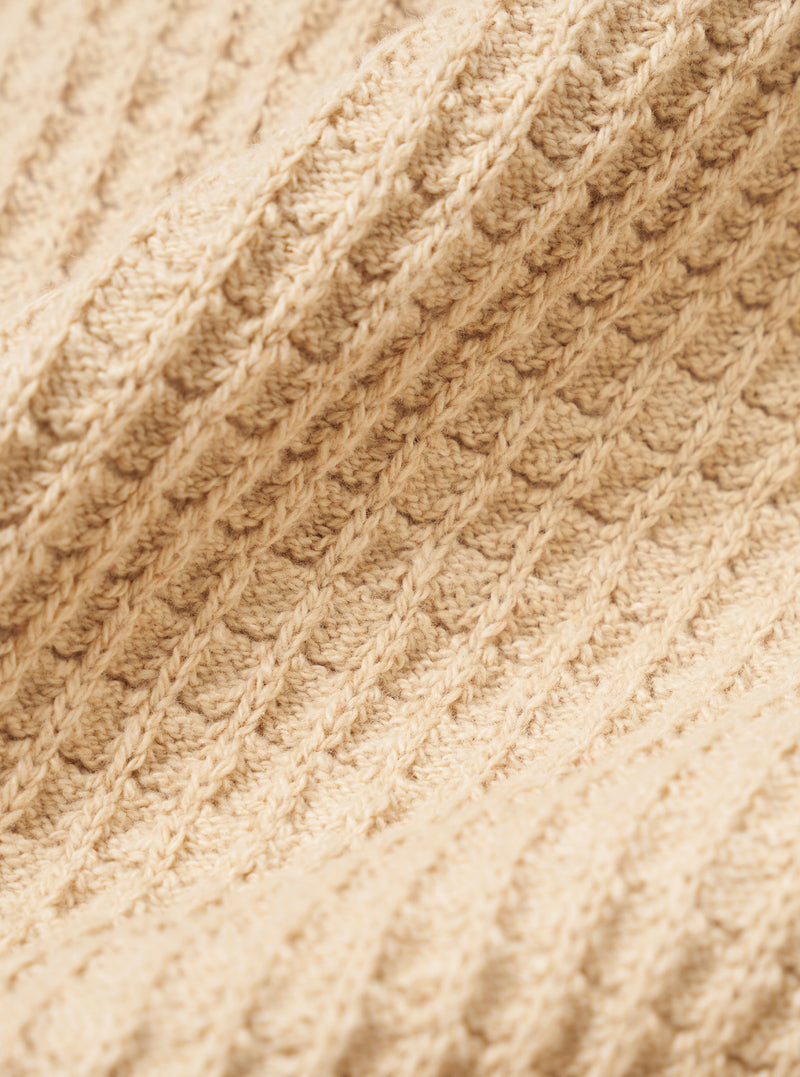 A close-up fabric swatch of the Sand Eco Cotton Knit Jacket shows the textured, ribbed knit of the recycled cotton fabric in detail. The sand-colored material fills the image, highlighting the weave and soft quality of the knitwear.