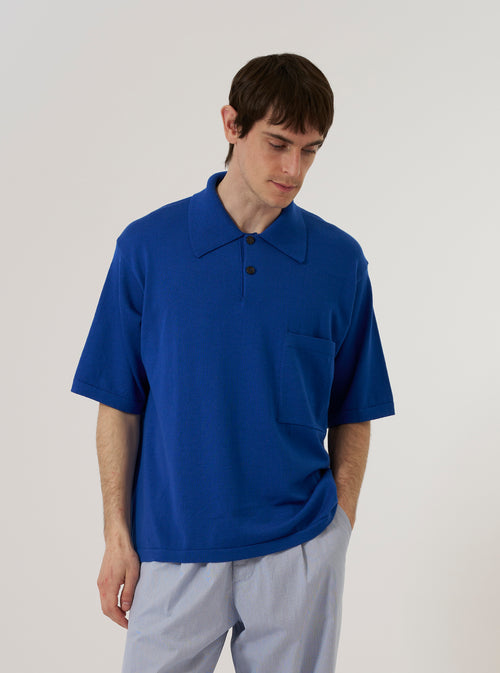 A model wears the Blue Woolcot Bio S/S Pullover Knit Shirt II against a plain, neutral background. The shirt features a short-sleeved pullover design with a button placket, a classic collar, and a single patch pocket on the left chest. The garment has a relaxed, boxy fit and is made from a smooth, dark blue knit fabric.