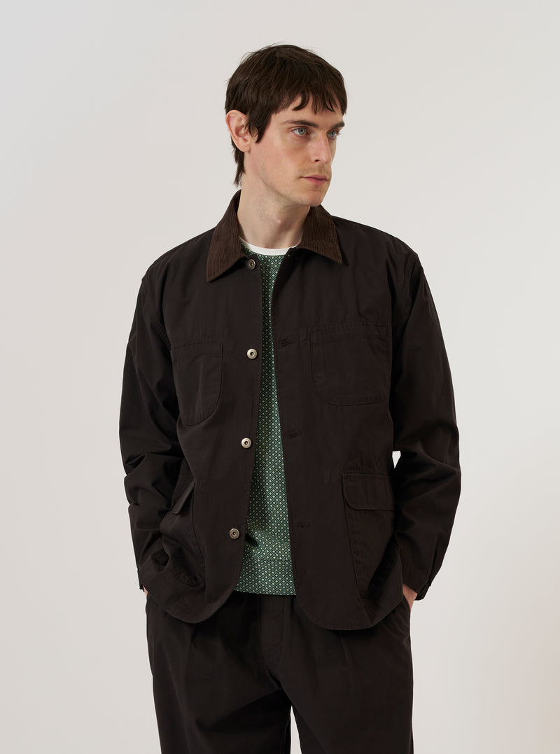 A model is wearing the Licorice Summer Canvas Labour Jacket over a light-colored top, standing against a plain white background. The jacket features a classic collar, a button-down front, two chest patch pockets, and two lower patch pockets with flap closures. The fabric is a solid brown licorice color with a sturdy canvas texture.