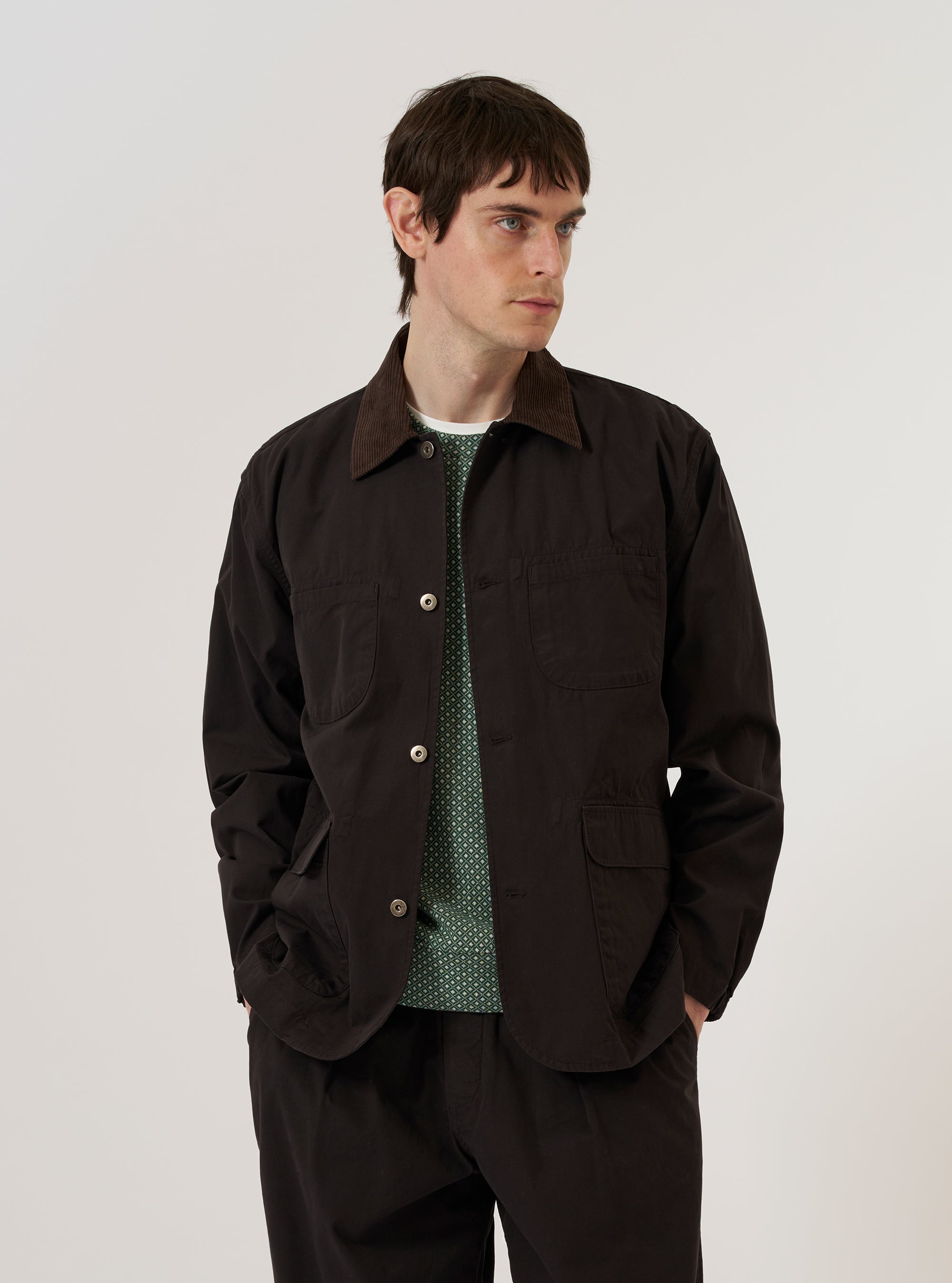A model is wearing the Licorice Summer Canvas Labour Jacket over a light-colored top, standing against a plain white background. The jacket features a classic collar, a button-down front, two chest patch pockets, and two lower patch pockets with flap closures. The fabric is a solid brown licorice color with a sturdy canvas texture.