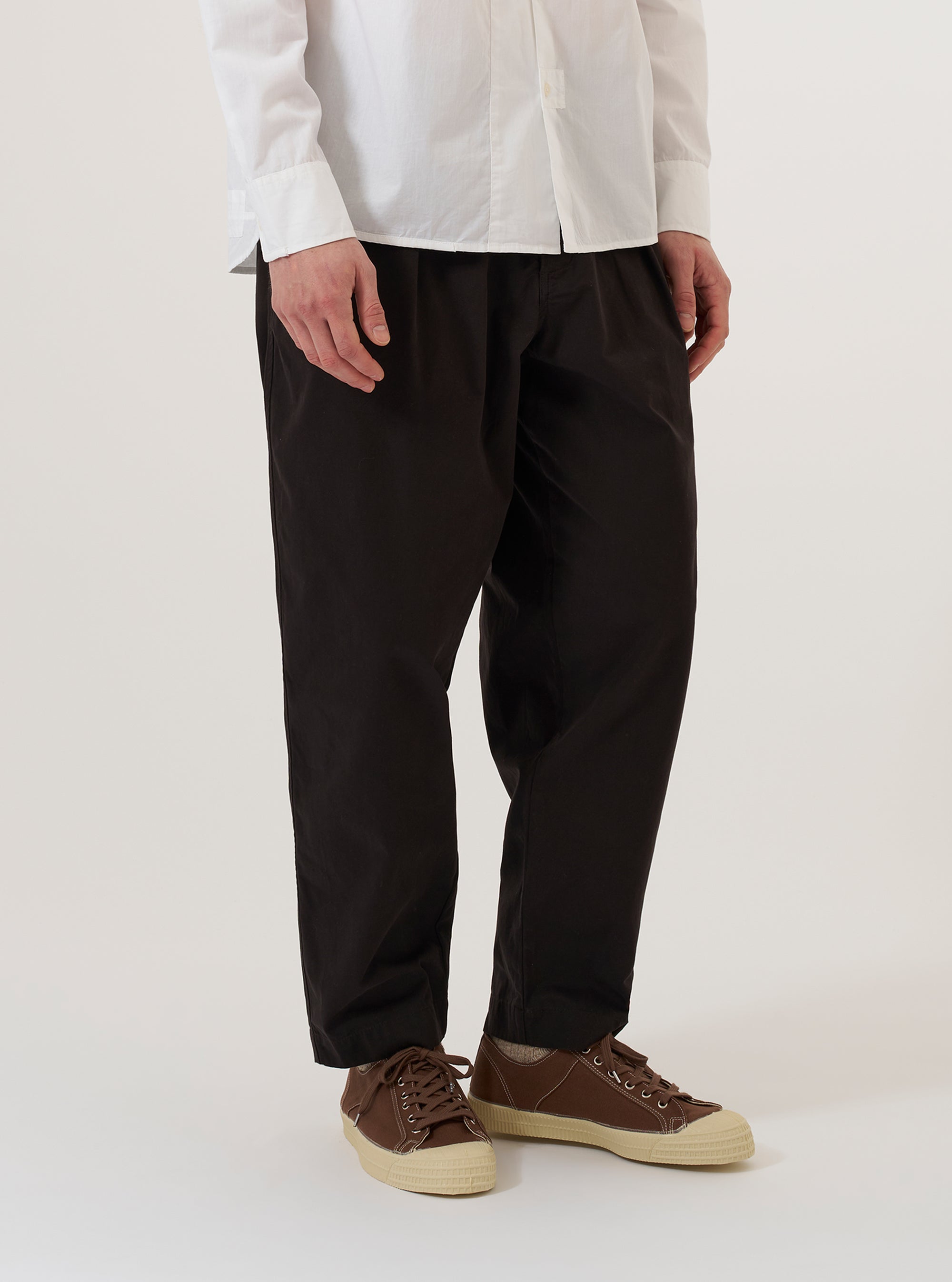 A model wears the Licorice Summer Canvas Pleated Track Pant against a plain white background. The image captures the trousers from the waist down, highlighting the elasticated waistband with an external drawstring and the prominent single pleats on the front of each leg. The garment is shown in a dark brown licorice color with a loose, tapered fit, styled with a light-colored top tucked in and dark shoes.