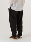 A model is wearing the Licorice Summer Canvas Pleated Track Pant, shown from the waist down from a side angle against a plain, neutral background. The dark brown trousers feature a loose, tapered fit, a pleated front, and an elasticated waistband with a functional drawstring. The summer canvas fabric has a smooth, durable texture.
