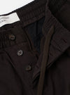 The Licorice Summer Canvas Pleated Track Pant is shown from the back as a flat lay against a plain white background. The image highlights the two rear pockets, one with a buttoned flap and one jetted with a button closure, along with the elasticated waistband and the textured, dark licorice-colored cotton canvas fabric.