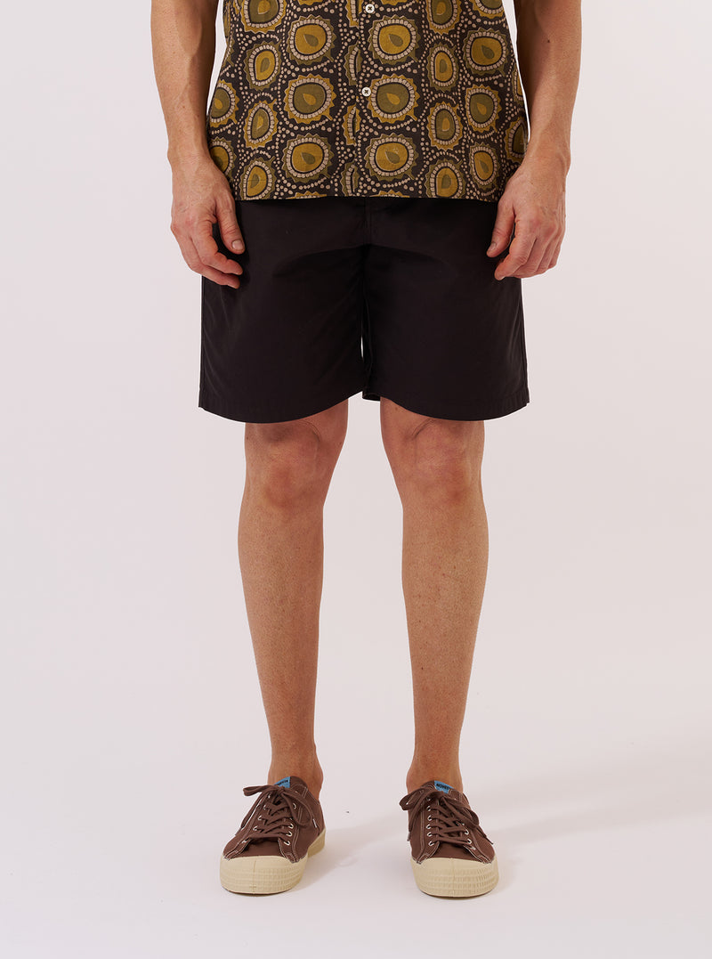 A model wears the Licorice Summer Canvas Clean Short, shown from the waist down against a plain white background. The shorts feature a relaxed fit, a mid-thigh length, and are made from a dark brown cotton canvas fabric. The model is wearing the shorts with white socks and brown shoes.