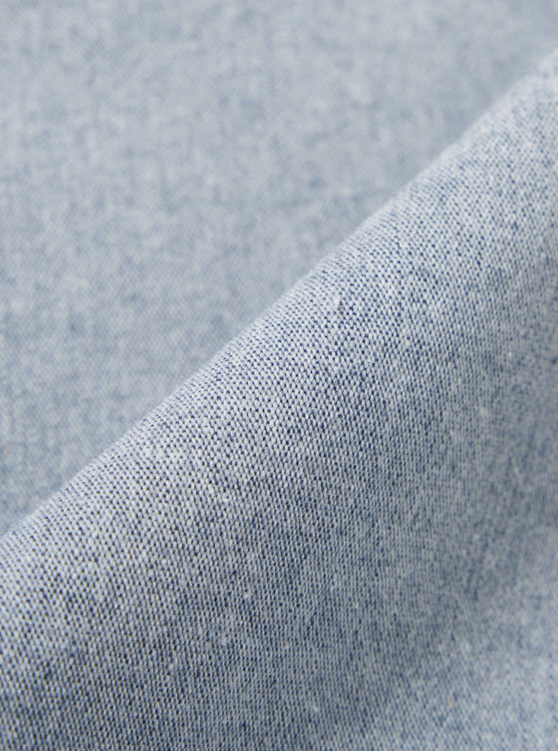 A close-up fabric swatch of the Light Indigo Jean Cloth Reworked Hi Water Trouser displays a light blue indigo denim with a subtle textured weave. The organic cotton fabric is shown in detail, filling the frame to highlight the material's color and construction.