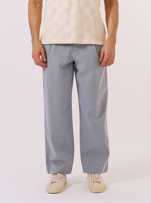 A model wears the Light Indigo Jean Cloth Reworked Duke Pant, shown from the waist down against a plain white background. The trousers feature a wide, straight leg with a single pleat at the front and a fixed waistband. The light indigo organic cotton fabric has a soft, washed appearance. The model is wearing the pants with white socks and brown shoes.
