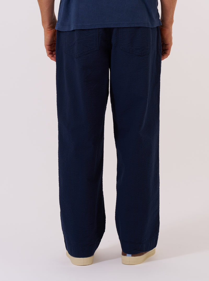 A model wears the Navy Derby Stripe Oxford Pant, shown from a rear three-quarter angle against a plain, neutral studio background. The trousers feature a loose, wide-leg fit and a navy and white vertical stripe pattern. The image highlights the clean back construction and the drape of the striped cotton fabric.