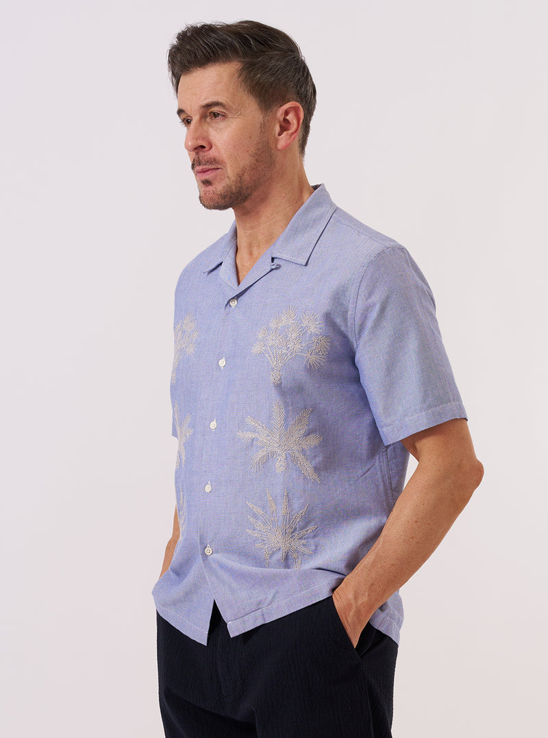 A model wears the Blue Oxford Shirting Road Trip Shirt while standing against a plain white background. The shirt features a camp collar, a single chest patch pocket, and a button-down front. It is worn open over a white t-shirt, showcasing the structured cotton oxford fabric and short sleeves.