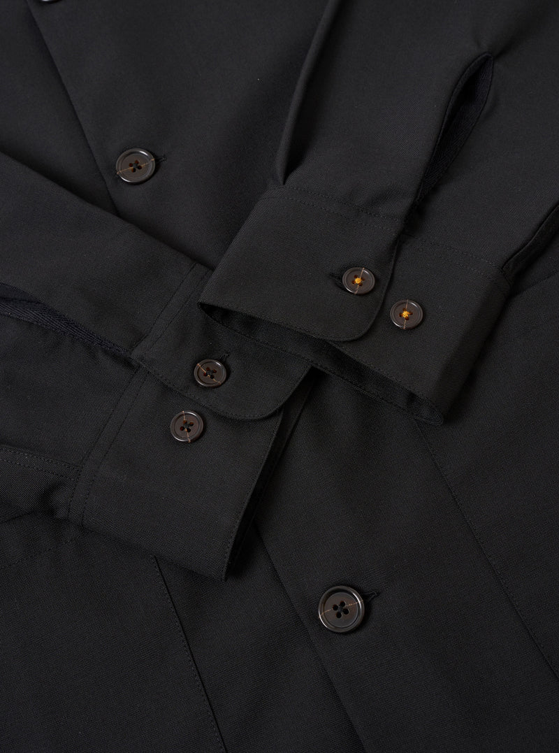 The Black Tropical Suiting Travail Overshirt is shown in a detail view as a flat lay against a plain white background. The image focuses on the lower front section of the garment, highlighting the smooth texture of the black tropical suiting fabric, the straight hem, and the clean stitching of a large patch pocket.