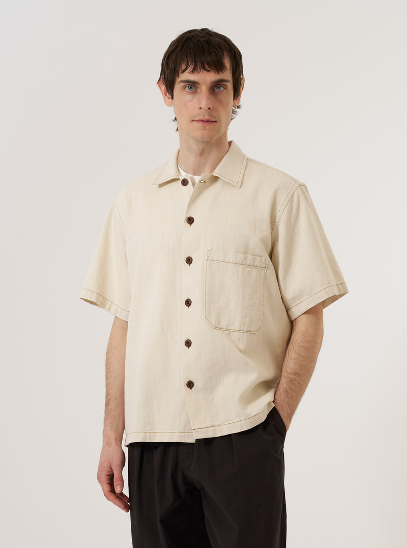 A model wears the Ecru Koko Cotlin S/S Fatigue Shirt standing against a plain white background. The shirt features a central button-down closure, a classic collar, and two prominent patch pockets on the chest. The short-sleeved garment is made from a light ecru fabric with a textured cotton-linen finish.