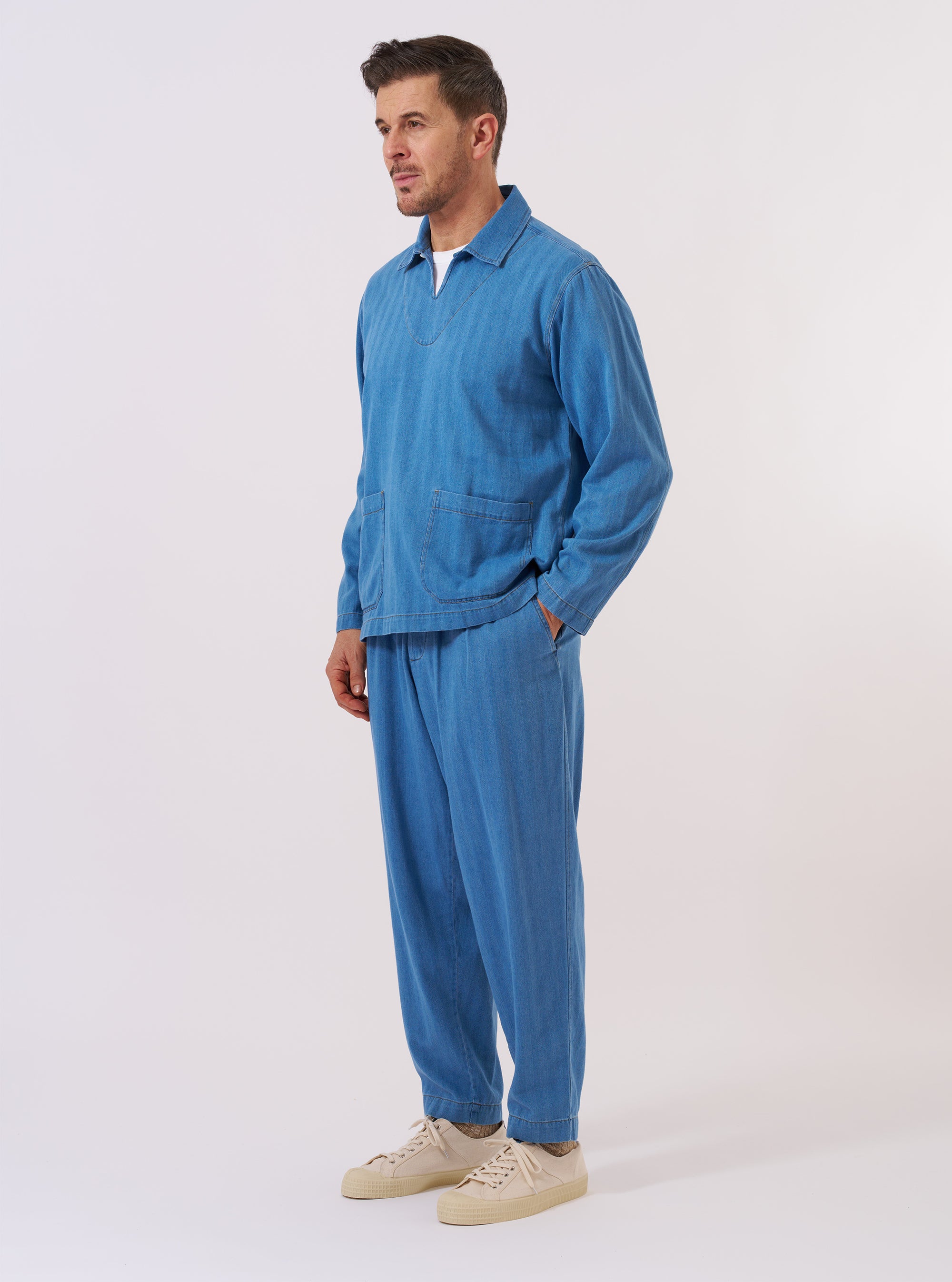 The Washed Indigo Recycled Herringbone Smock Overshirt is shown as a flat lay against a plain white background. The pullover-style garment features a partial button-down front with three visible buttons, a classic pointed collar, and a single patch pocket on the left chest. The washed indigo blue fabric displays a distinct herringbone weave pattern.