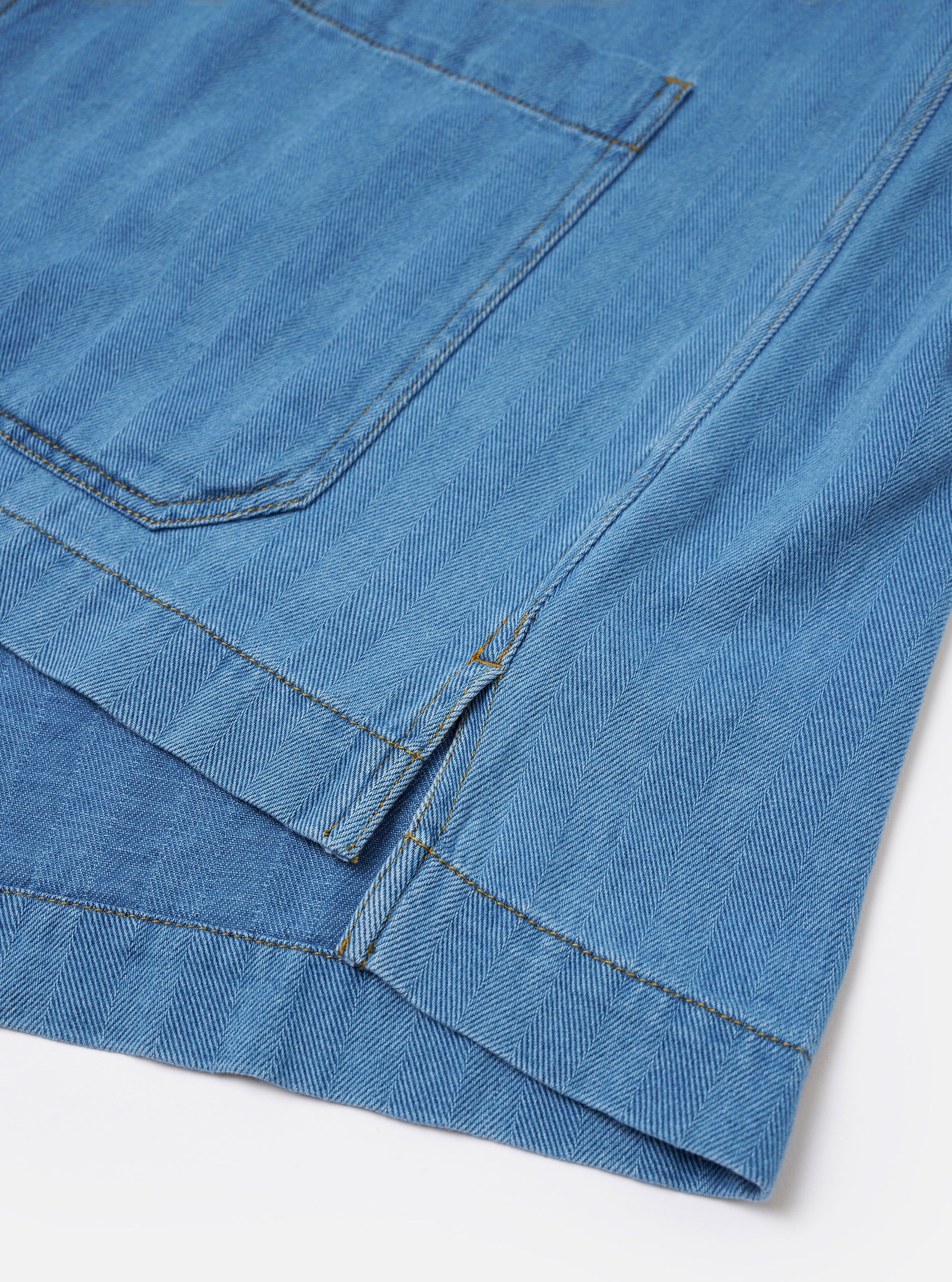 The Washed Indigo Recycled Herringbone Smock Overshirt is shown from the back as a flat lay against a plain white background. The image highlights the garment's textured herringbone weave and the deep washed indigo blue color of the recycled cotton fabric. The back features a simple, clean construction with a straight hem and visible tonal stitching along the seams.