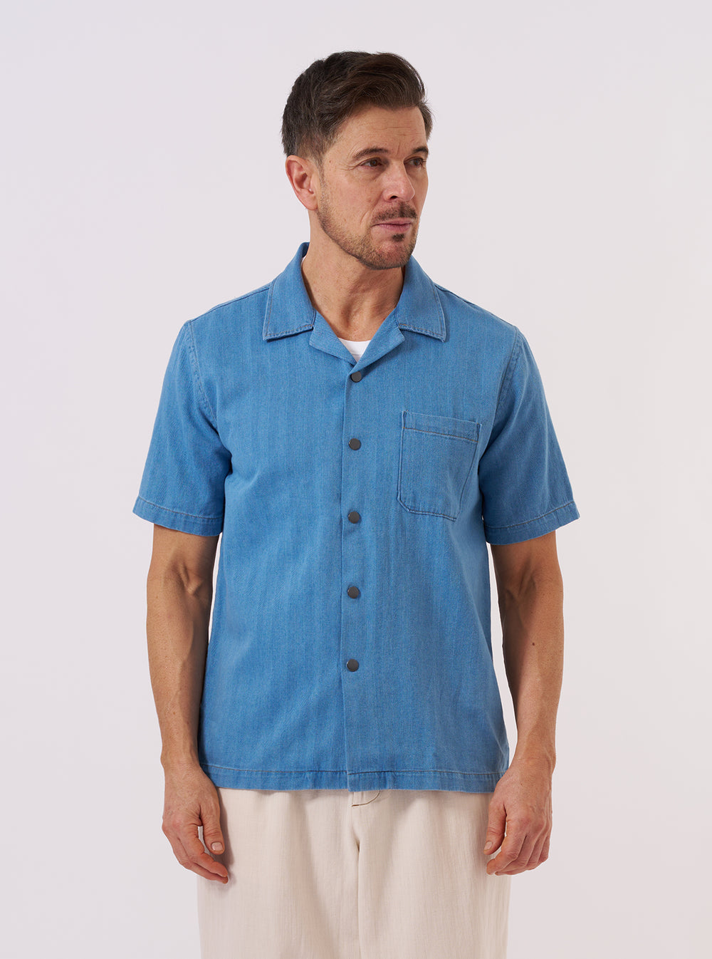 A model wears the Washed Indigo Recycled Herringbone Road Shirt while standing against a plain, neutral background. The shirt features a relaxed fit with a camp collar and a central button-down front. The washed indigo fabric is characterized by a visible herringbone weave pattern throughout the garment.