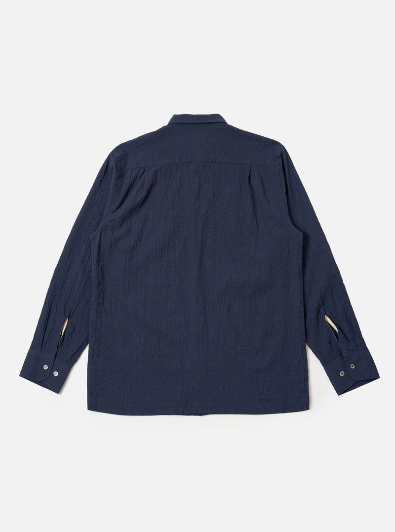 The Navy Seersucker Shirting Lucky Shirt is shown from the back as a flat lay against a plain white background. The image highlights the shirt's relaxed fit, straight hem, and the subtle puckered texture of the navy blue seersucker fabric. A simple back yoke and clean stitching along the hem and sleeves are visible.